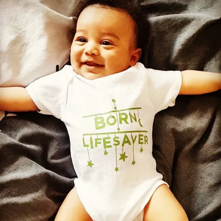 Smiling baby in a &lsquo;Born lifesaver&rsquo; babygrow with arms outstretched