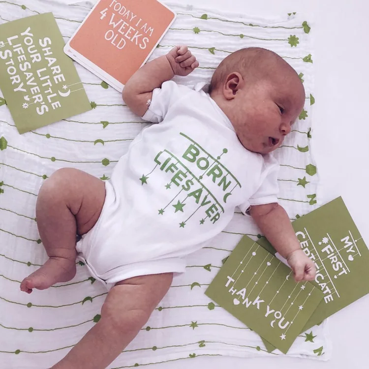 Sleeping newborn in a &lsquo;Born lifesaver&rsquo; babygrow with a &lsquo;4 weeks old&rsquo; card, postcards and certificate
