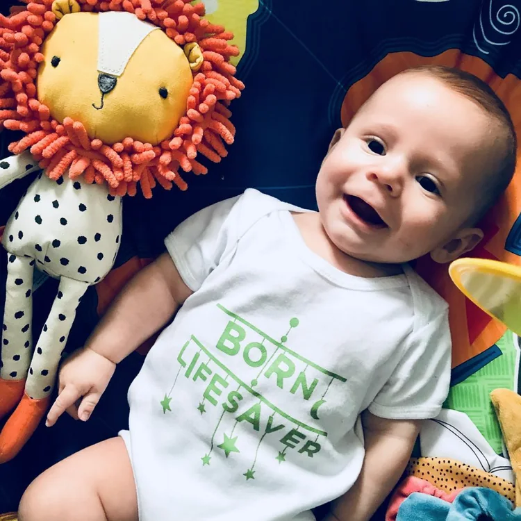 Smiling baby in a &lsquo;Born lifesaver&rsquo; babygrow beside a toy lion