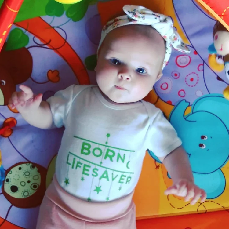 Baby with a floral headband in a &lsquo;Born lifesaver&rsquo; babygrow on a play mat