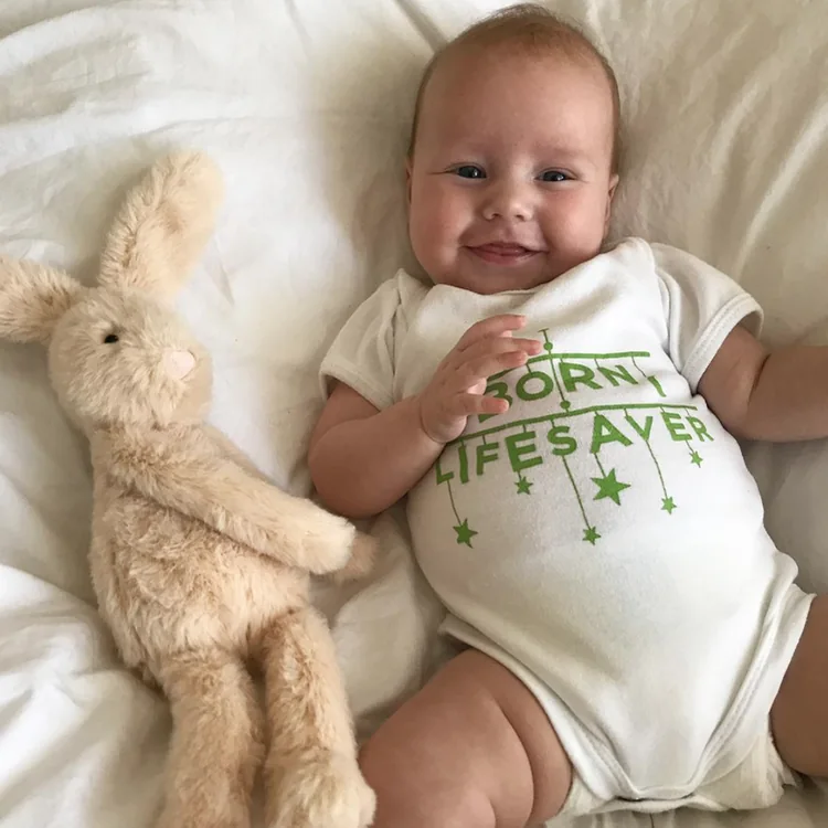 Smiling baby in a &lsquo;Born lifesaver&rsquo; babygrow next to a toy rabbit