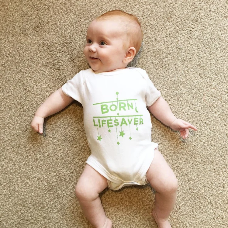Smiling baby in a &lsquo;Born lifesaver&rsquo; babygrow lying on a carpet