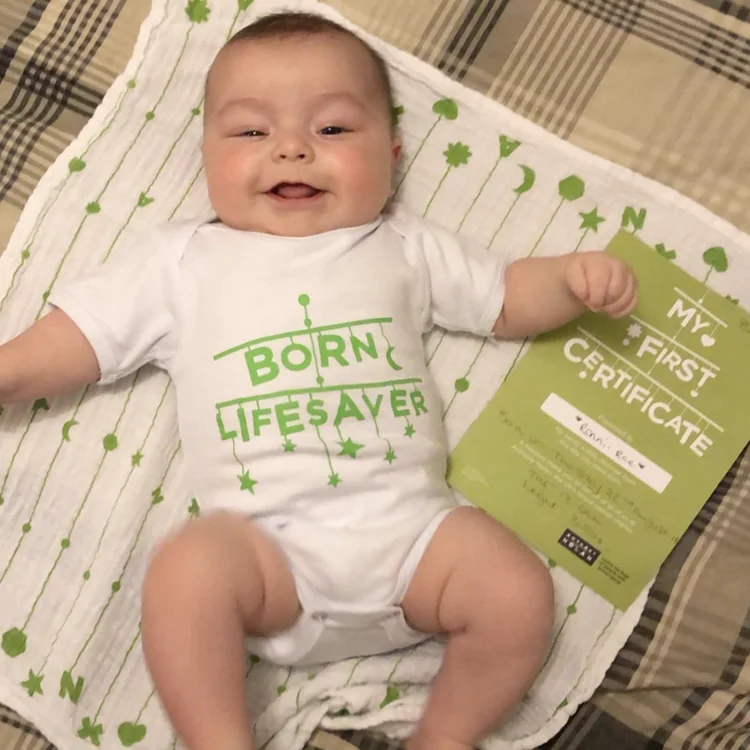 Smiling baby on the muslin in a &lsquo;Born lifesaver&rsquo; babygrow with the certificate