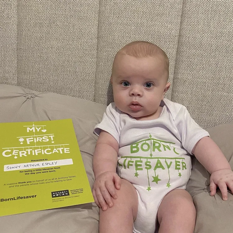 Baby in a &lsquo;Born lifesaver&rsquo; babygrow sitting up beside the certificate
