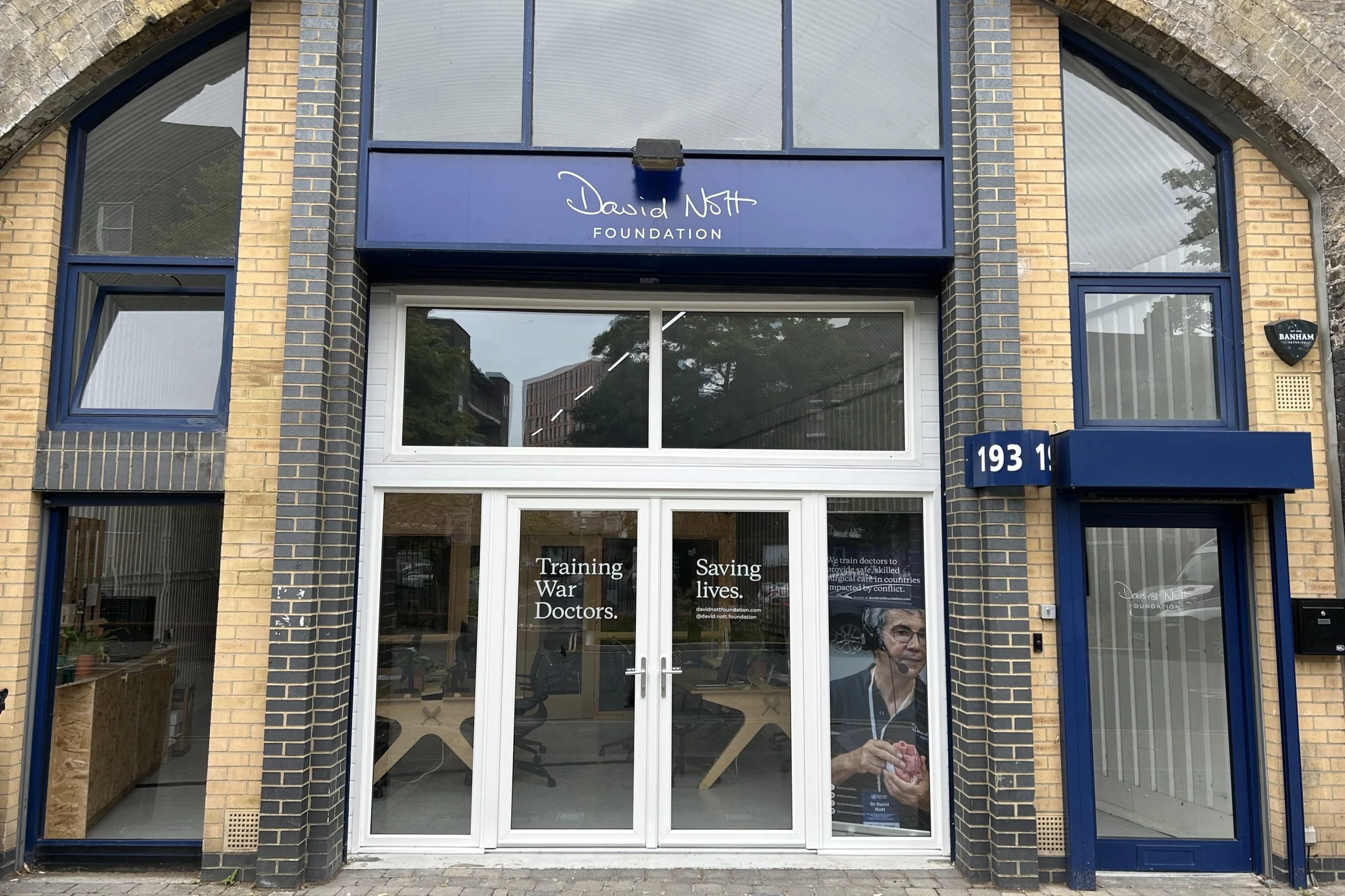 The David Nott Foundation office in a railway arch, with the signature logo on a blue fascia and &lsquo;Training war doctors. Saving lives.&rsquo; vinyls on the glass doors