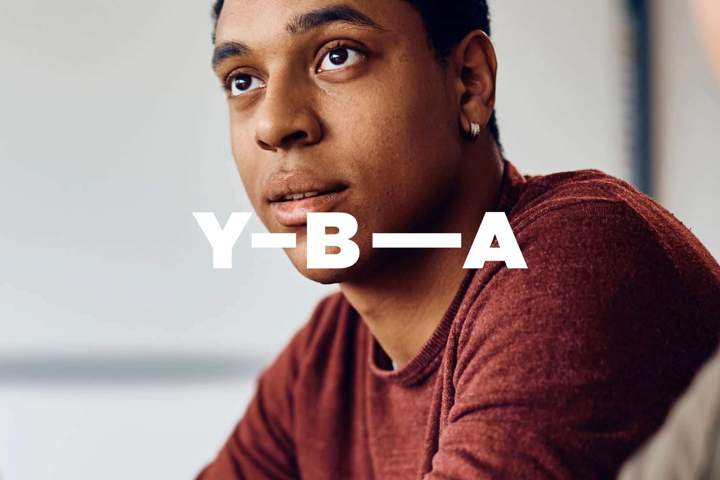 Photographic style: a young man looking thoughtfully into the distance, with the Y-B-A wordmark overlaid in white