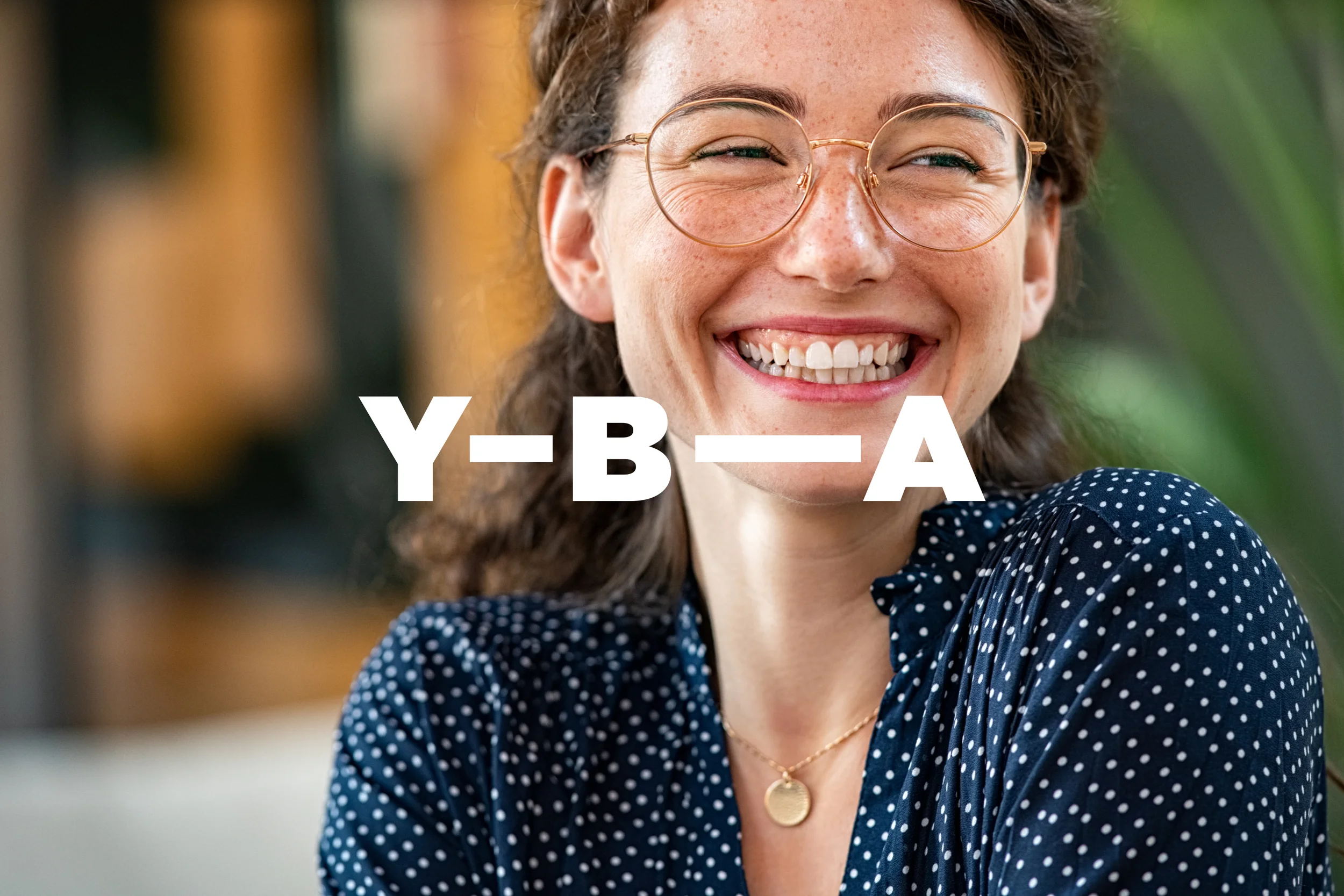 Photographic style: a young woman in glasses laughing, with the Y-B-A wordmark overlaid in white